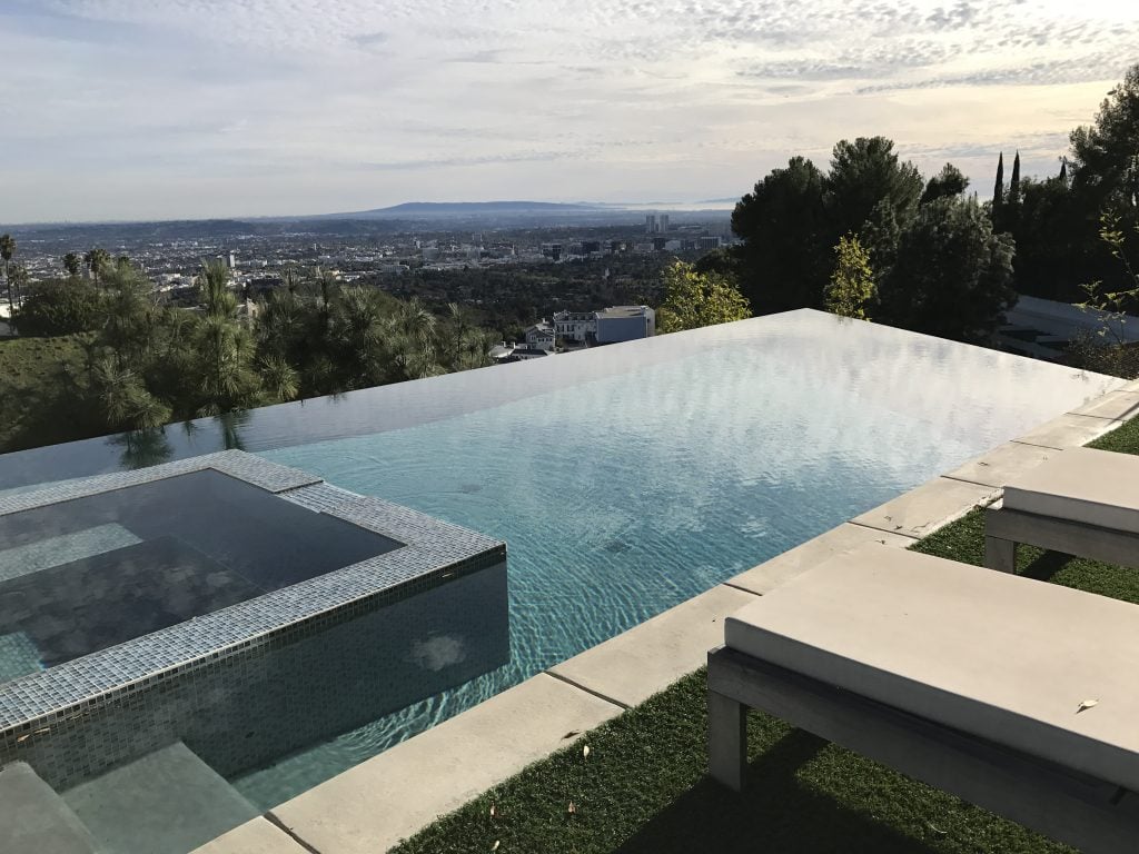 Modern infinity pool in the hills of Beverly Hills with city views stretching toward Los Angeles under a partly cloudy sky.