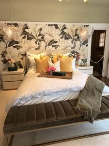 Styled bedroom interior with layered bedding, patterned wallpaper, and warm natural lighting.